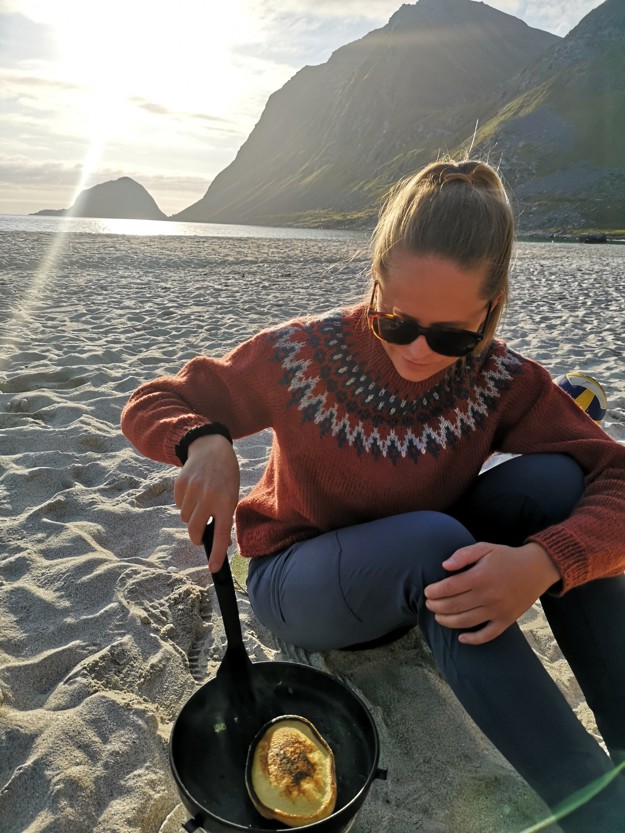 Astrid on a beach with a mountain behind her. She is wearing a wool sweater and is frying a pancake in a frying pan.