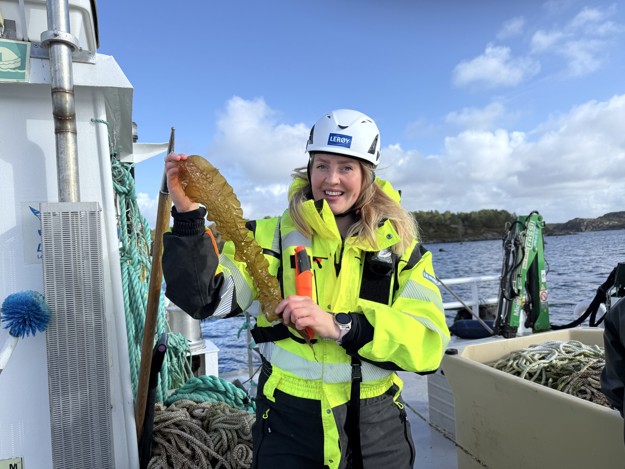 A woman in bright yellow work-wear, she is holding up a pice of a kelp and is smiling, looking at the camera. 