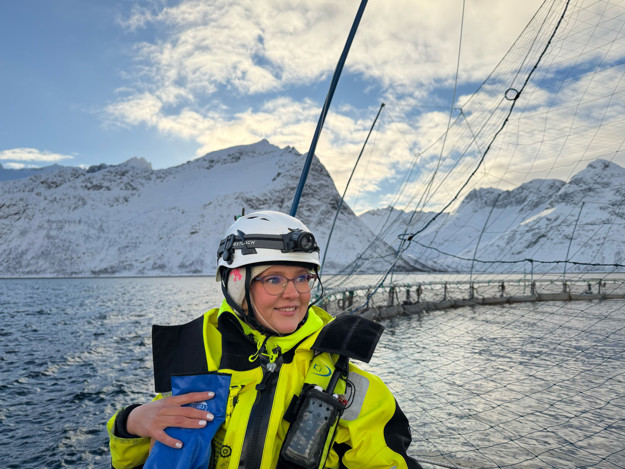 Ung kvinne på sjøen med arbeidsklær, bak henne er sjøen med en merd og fjell